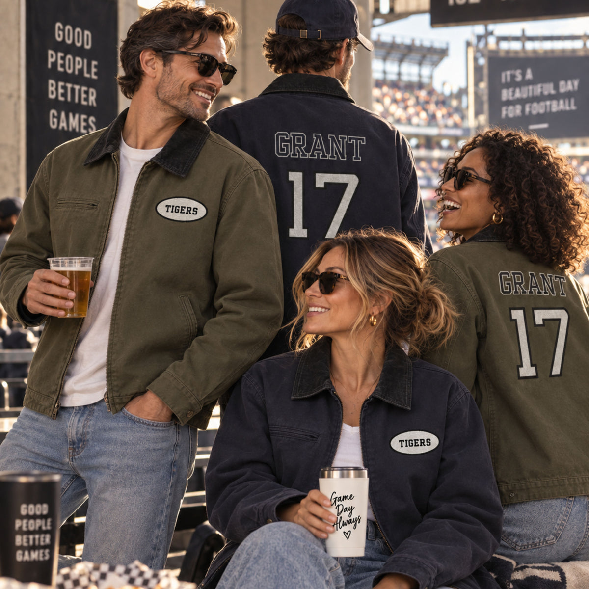 Four people wearing matching sports jackets in a stadium setting. Two jackets are olive green with black collar panels and a white oval 'Tigers' patch, while two are navy blue with black collar panels and a white oval patch.