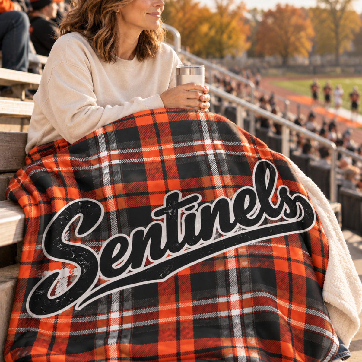 A person sitting in stadium seating at a sports field wrapped in a black, and orange plaid blanket featuring the word 'Sentinels' in large black script with a white outline.