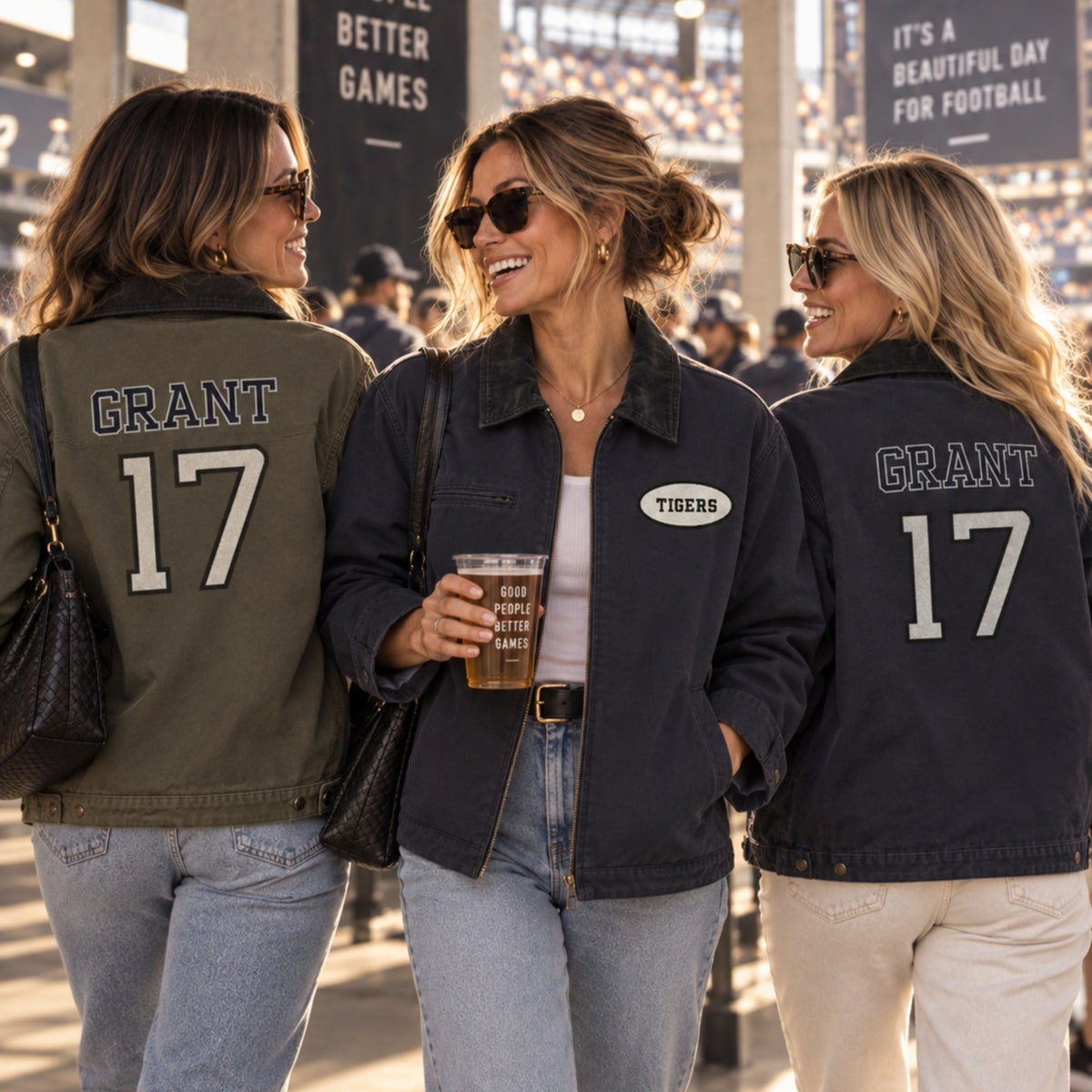 Three women walking together in a stadium, each wearing a customized number 17 sports jacket. The woman on the left wears an olive green jacket with 'GRANT' and '17' on the back, the center woman wears a navy blue jacket with a 'TIGERS' crest patch, and the woman on the right wears a navy blue jacket with 'GRANT' and '17' on the back.