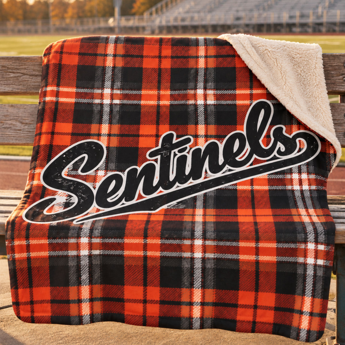 A fleece blanket draped over a wooden bench outdoors, featuring a orange, black, and white plaid pattern with a large 'Sentinels' script logo across the front and a cream sherpa lining visible on the folded top corner.
