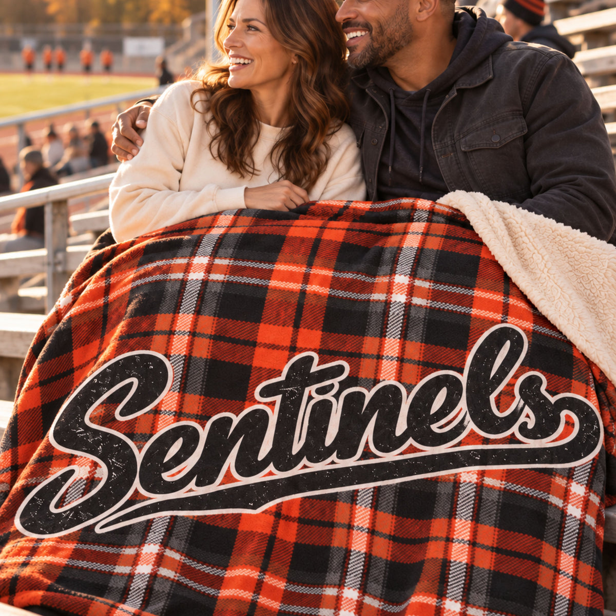 A couple sitting together in outdoor stadium seating, wrapped in a black, and orange plaid blanket with a cream sherpa side and a large 'Sentins' logo across the front.