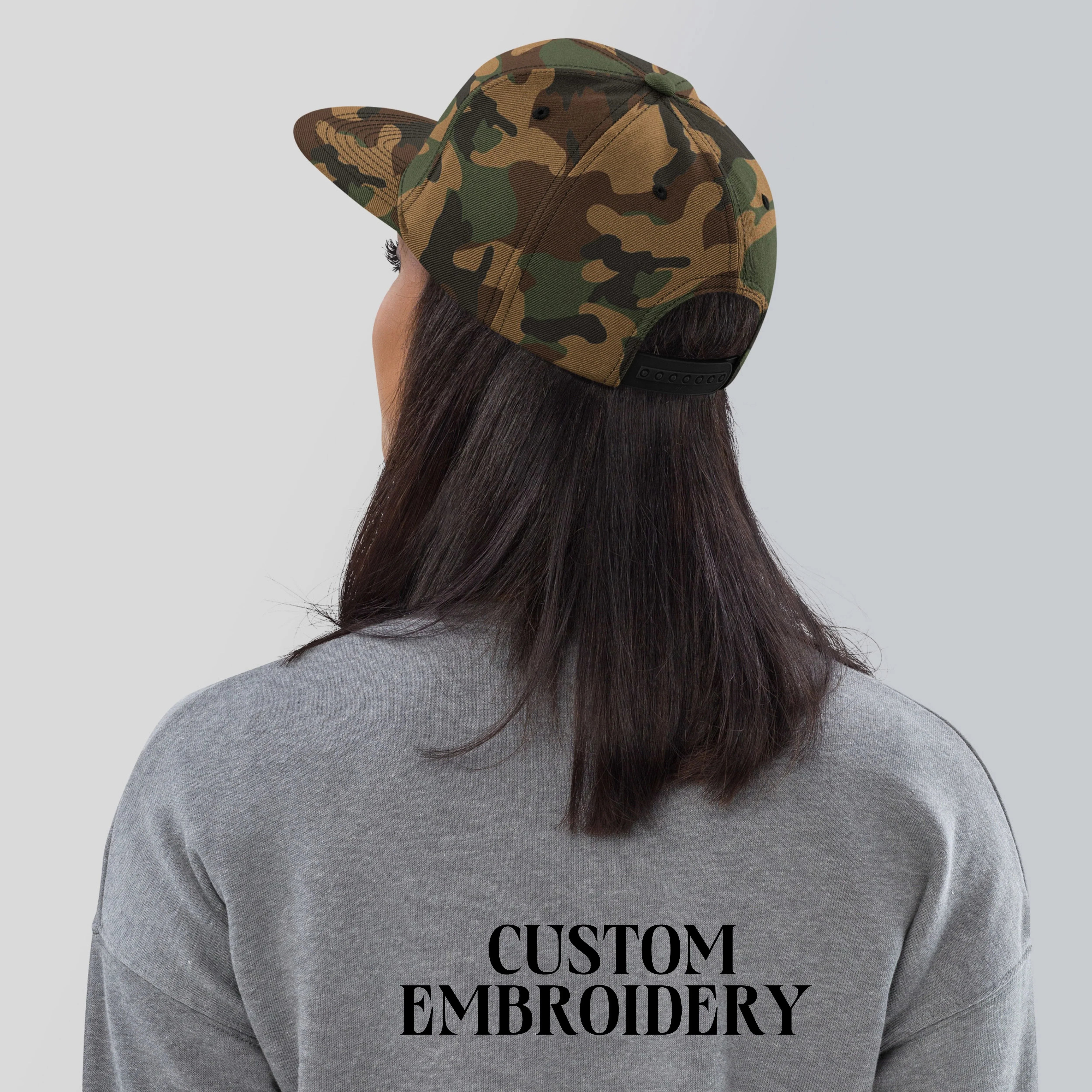 Woman wearing camo snapback hat and grey sweatshirt with custom embroidery on back