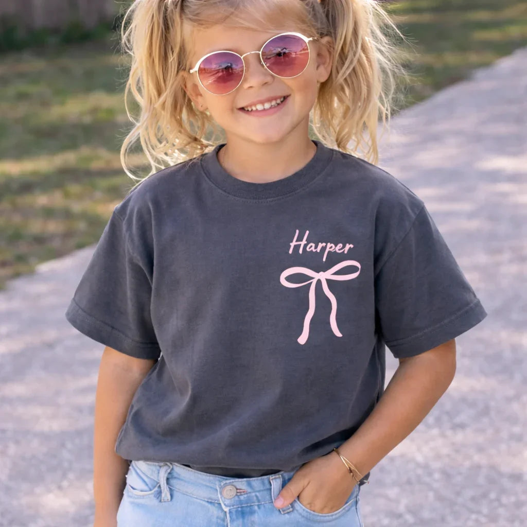Child wearing a dark gray t-shirt with 'Custom Name' and a pink bow design, standing outdoors. God Says I Am