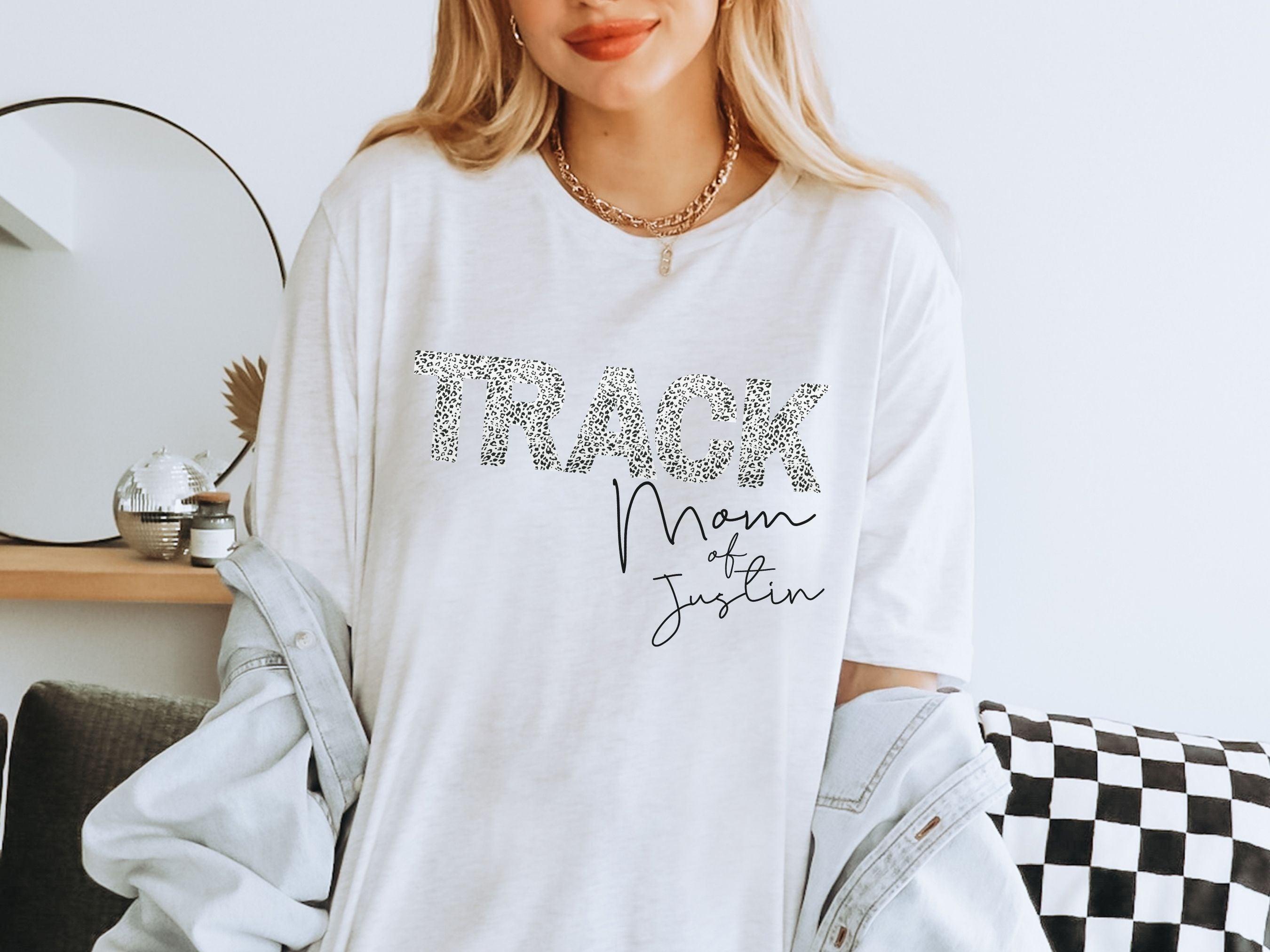 Woman wearing a white sweatshirt with 'Track Mom of Justin' text, sitting on a checkered couch.