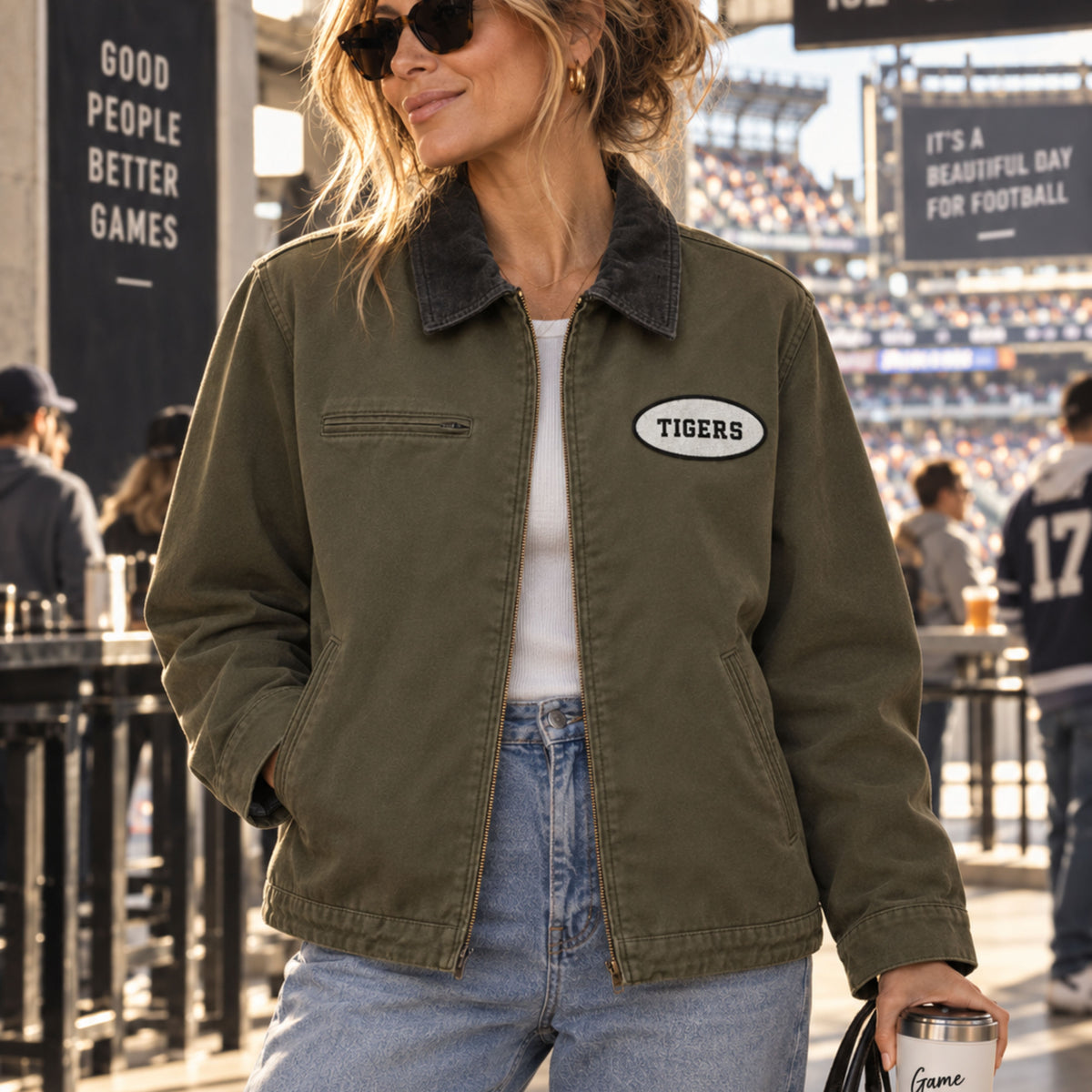A woman wearing an olive green utility jacket with a dark corduroy collar and a white 'Tigers' patch on the chest, standing in a stadium crowd.