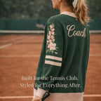 Woman on tennis court wearing green sweater with floral sleeve and 'Coach' text on back