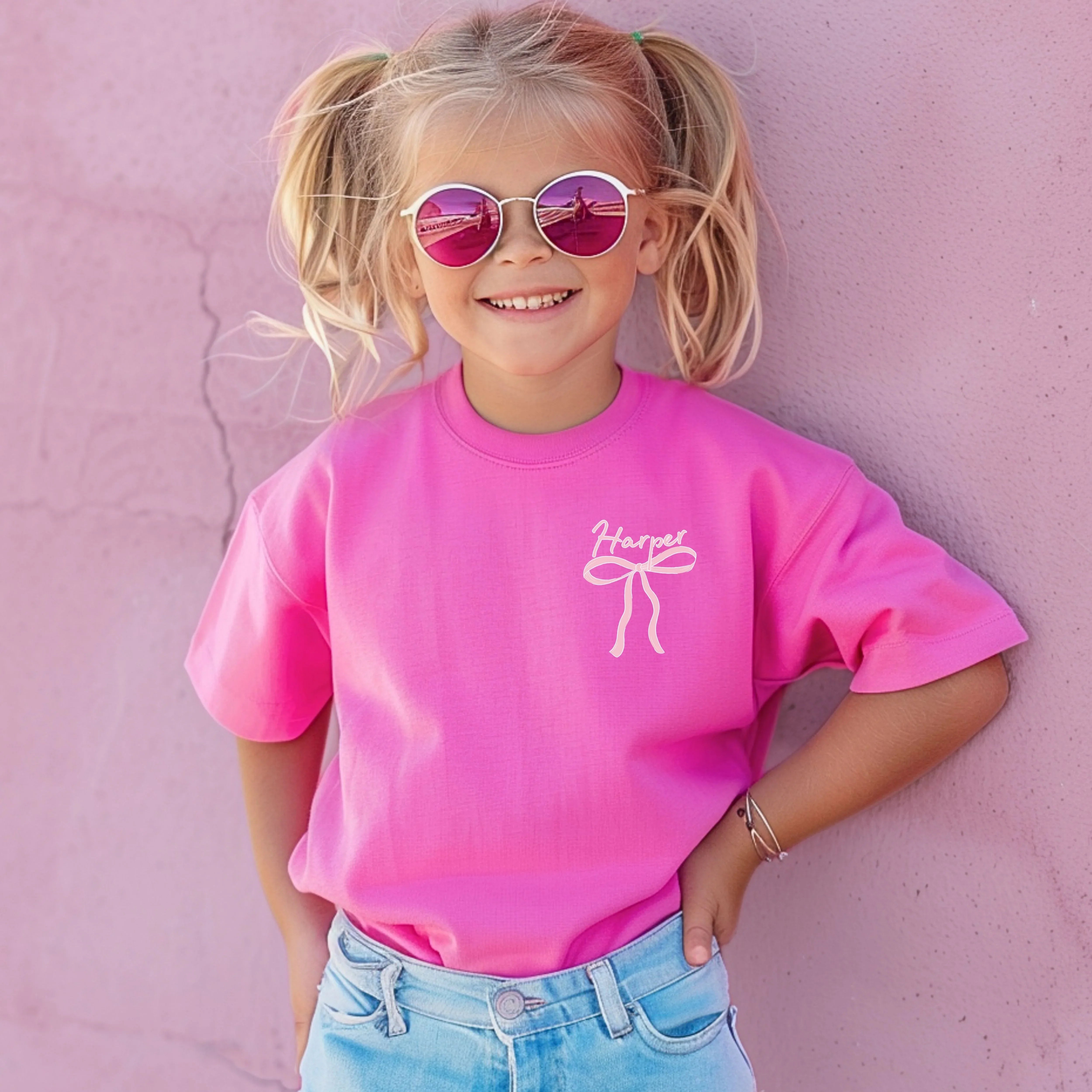 Child wearing a pink t-shirt with a custom name above a pink bow against a pink background