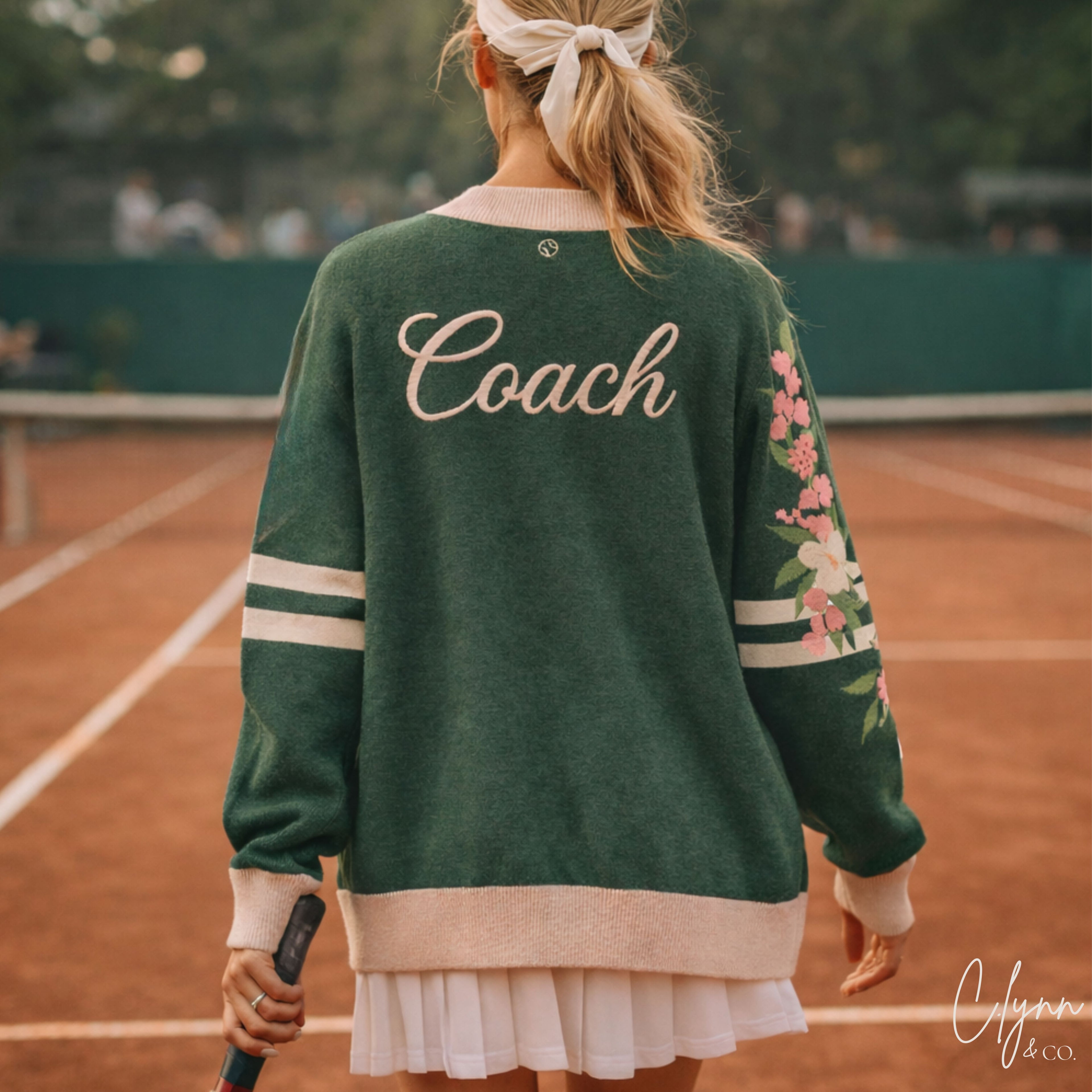 Woman in green Coach sweater with floral sleeve holding tennis racket on clay court
