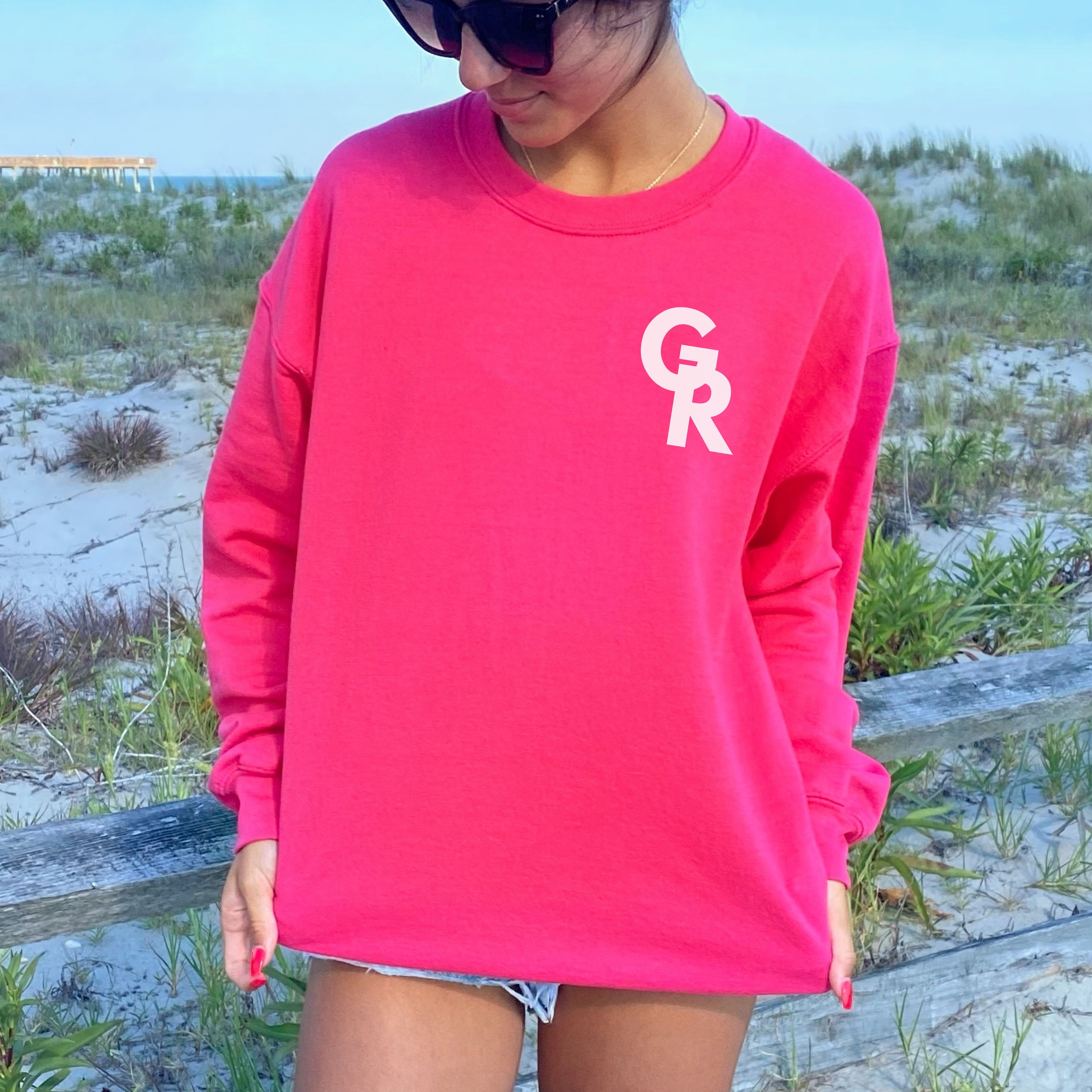 Person wearing a bright pink sweatshirt with 'GR' logo on a beach Gym Rat