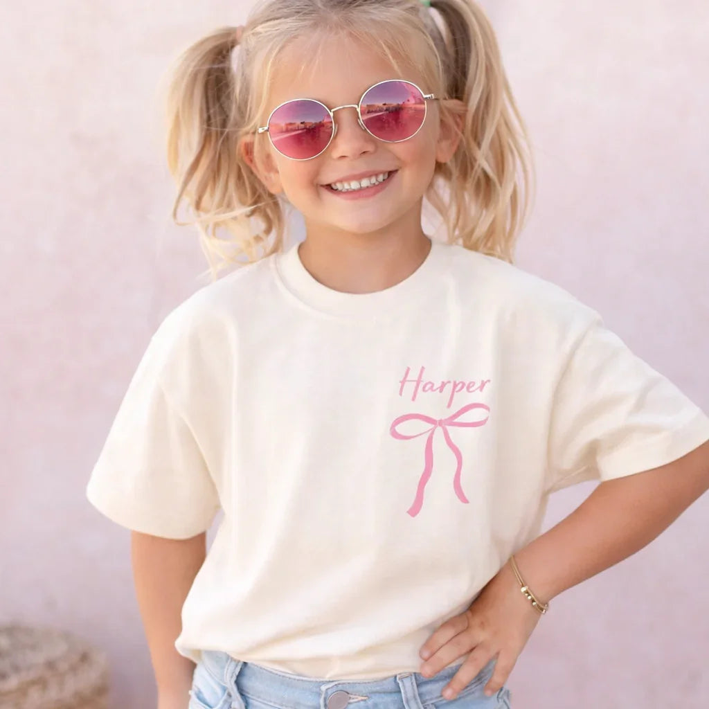 Child wearing a white t-shirt with a pink bow and name 'Harper' on a light background