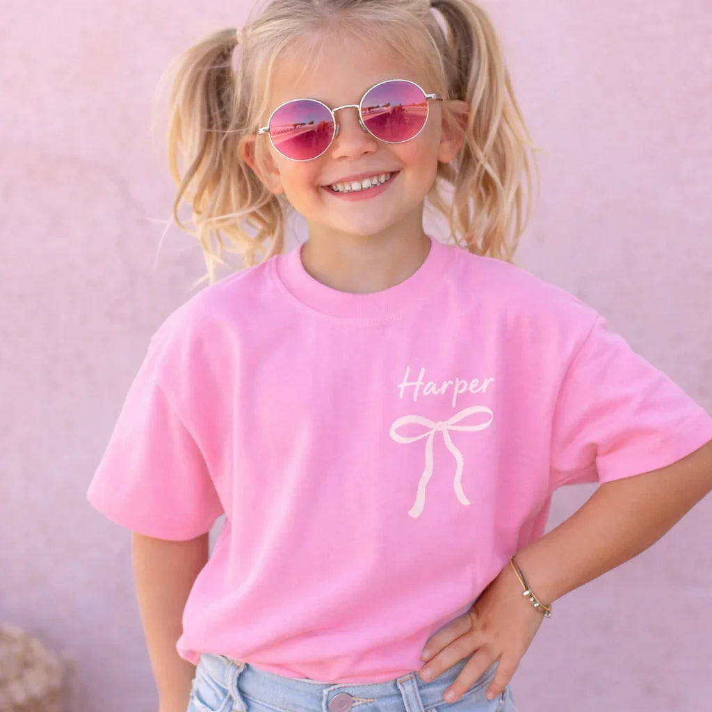 Child wearing a pink t-shirt with a bow design and name 'Harper' on a pink background