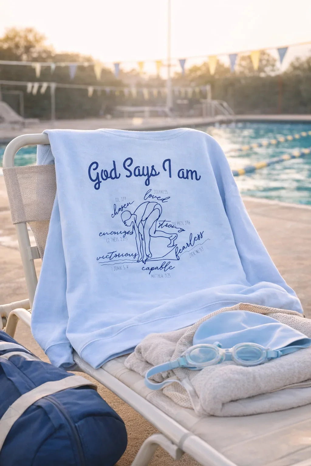 Light blue God Says I am Swimmer sweatshirt with text and graphics on a lounge chair by a pool.