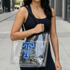 Woman holding a clear bag with a logo, wearing a black cap and tank top, on a city street.