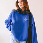 Person wearing a blue sweatshirt with a Heart and Cross symbol on the chest pocket area of the shirt, holding black headphones, on a light pink background