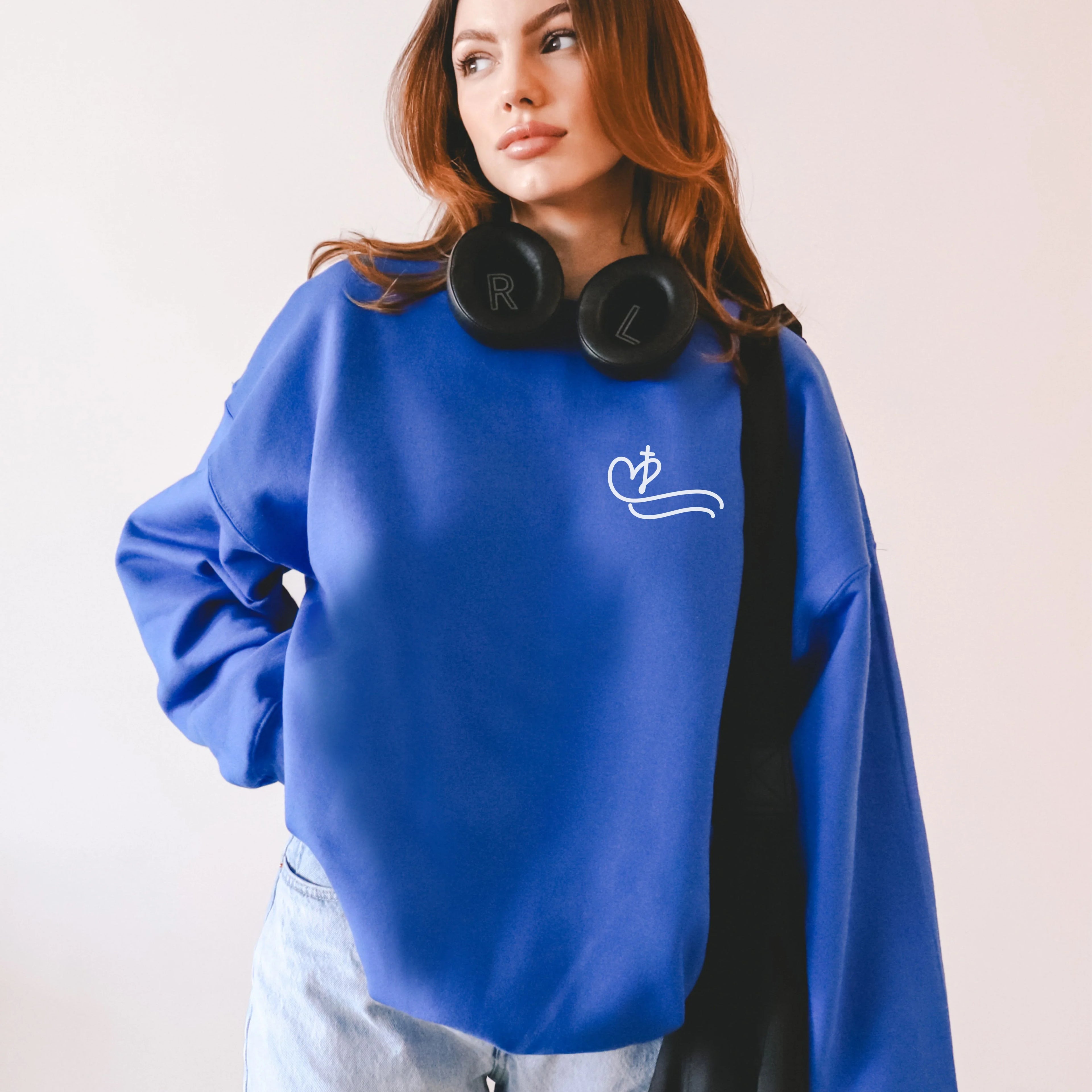 Person wearing a blue sweatshirt with a Heart and Cross symbol on the chest pocket area of the shirt, holding black headphones, on a light pink background