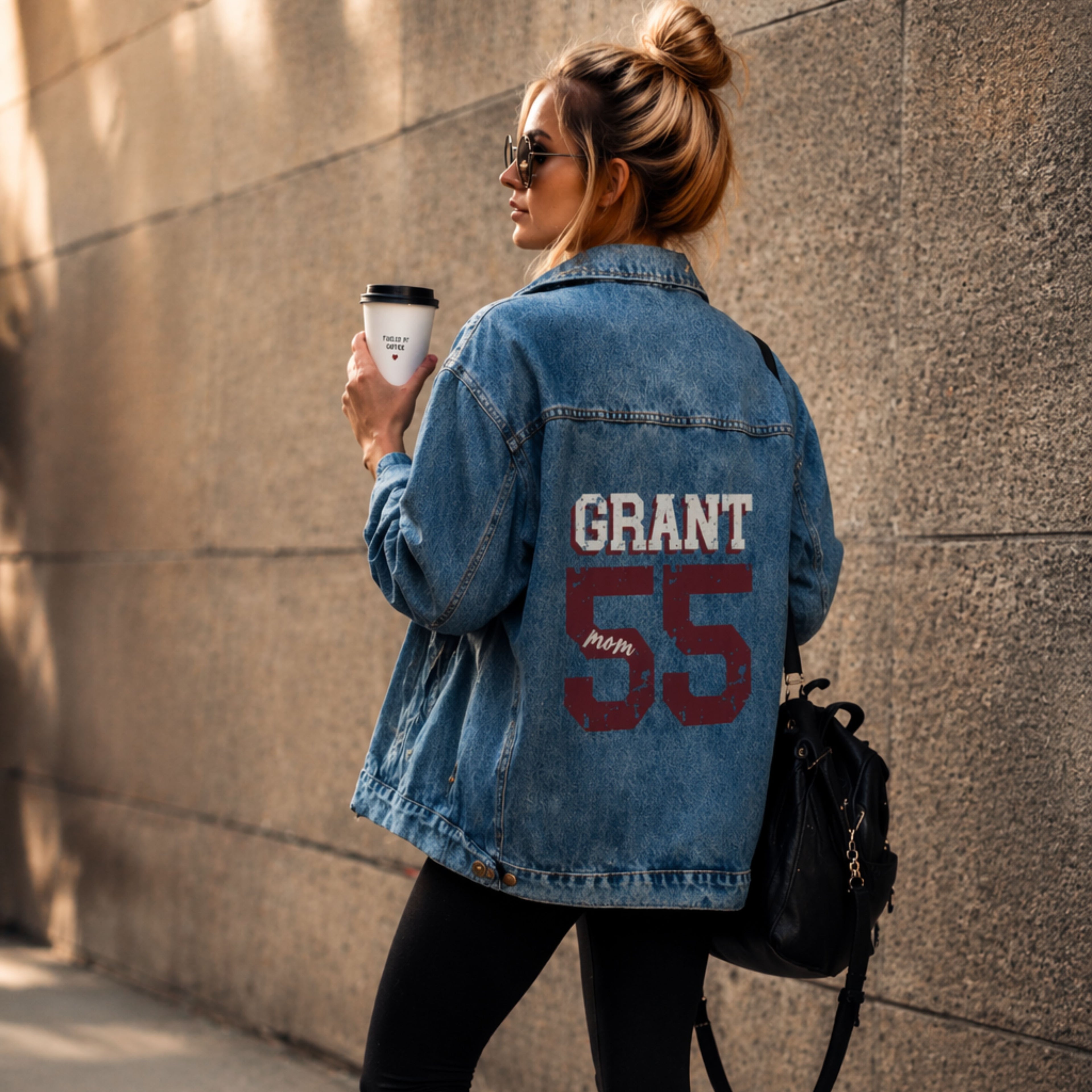 Woman wearing a denim jacket with 'GRANT 55' on it, holding a coffee cup against a wall.