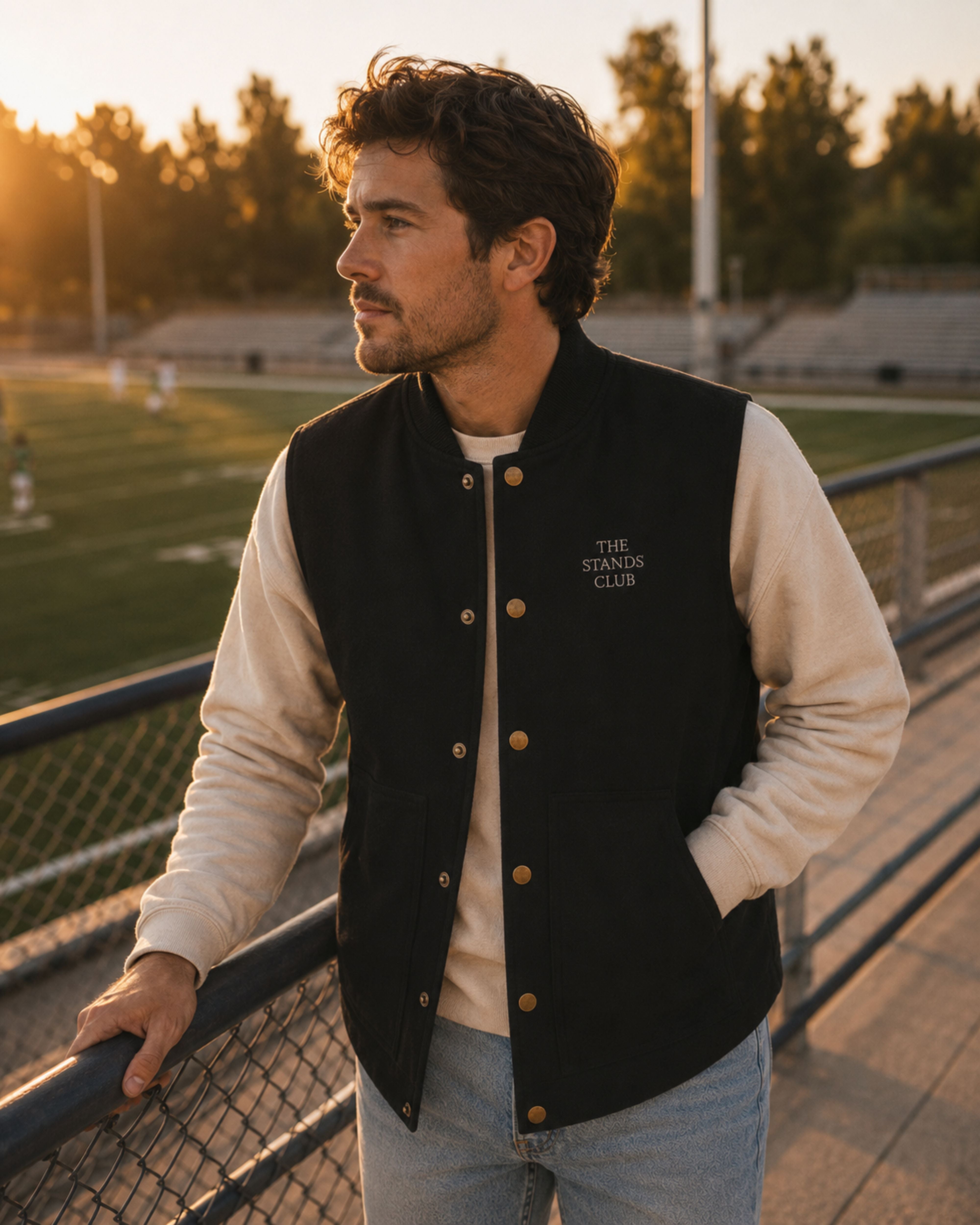 A man wearing a varsity-style jacket with black body and cream sleeves, standing on a walkway at a sports field during golden hour. The jacket has gold-tone buttons and 'THE STANLEY CLUB' embroidered on the left chest.
