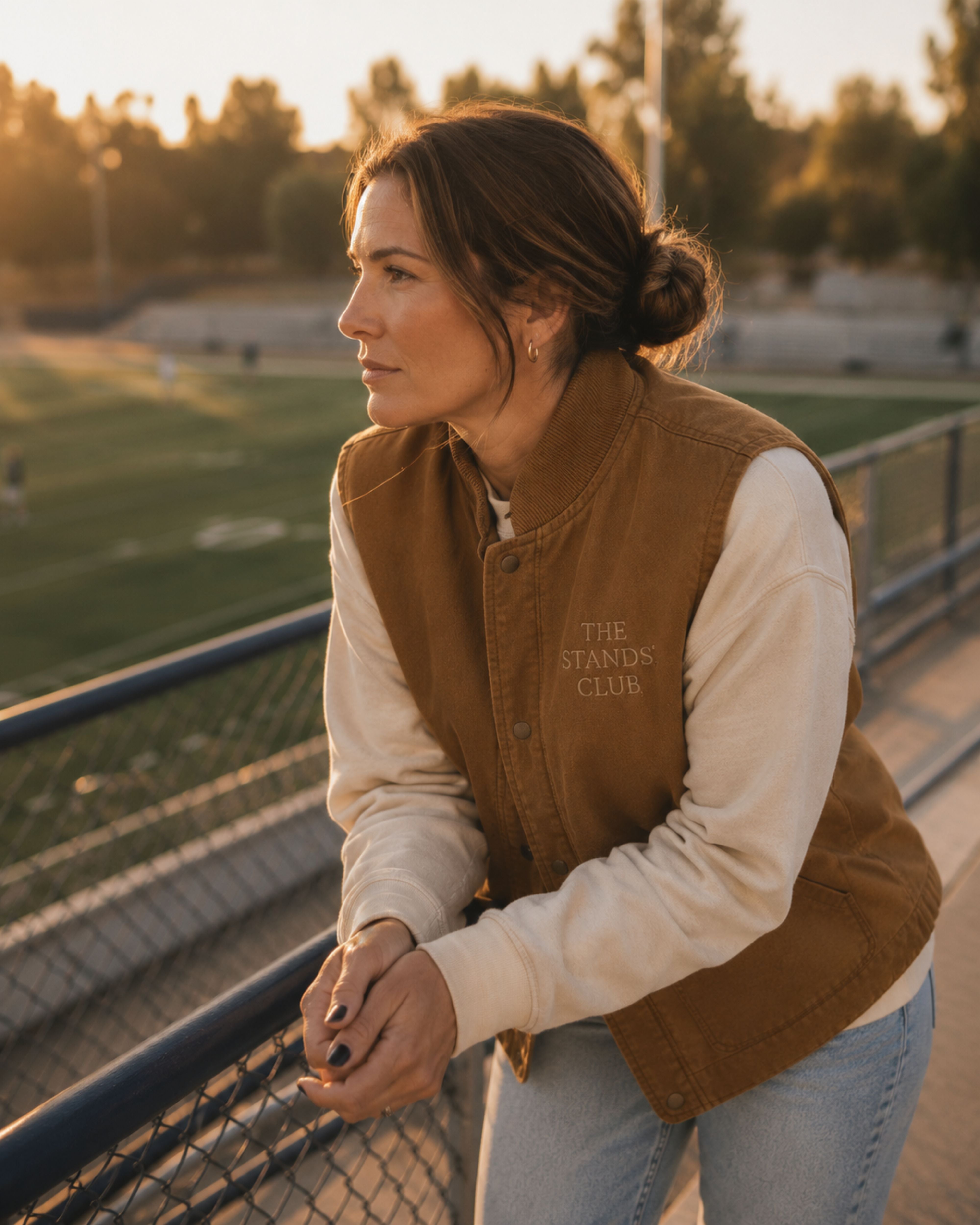 A woman stands at a metal railing at an outdoor sports field during golden hour, wearing a brown varsity-style jacket over a cream long-sleeve shirt and light blue jeans. The jacket has 'THE STANDS CLUB' embroidered on the left chest.