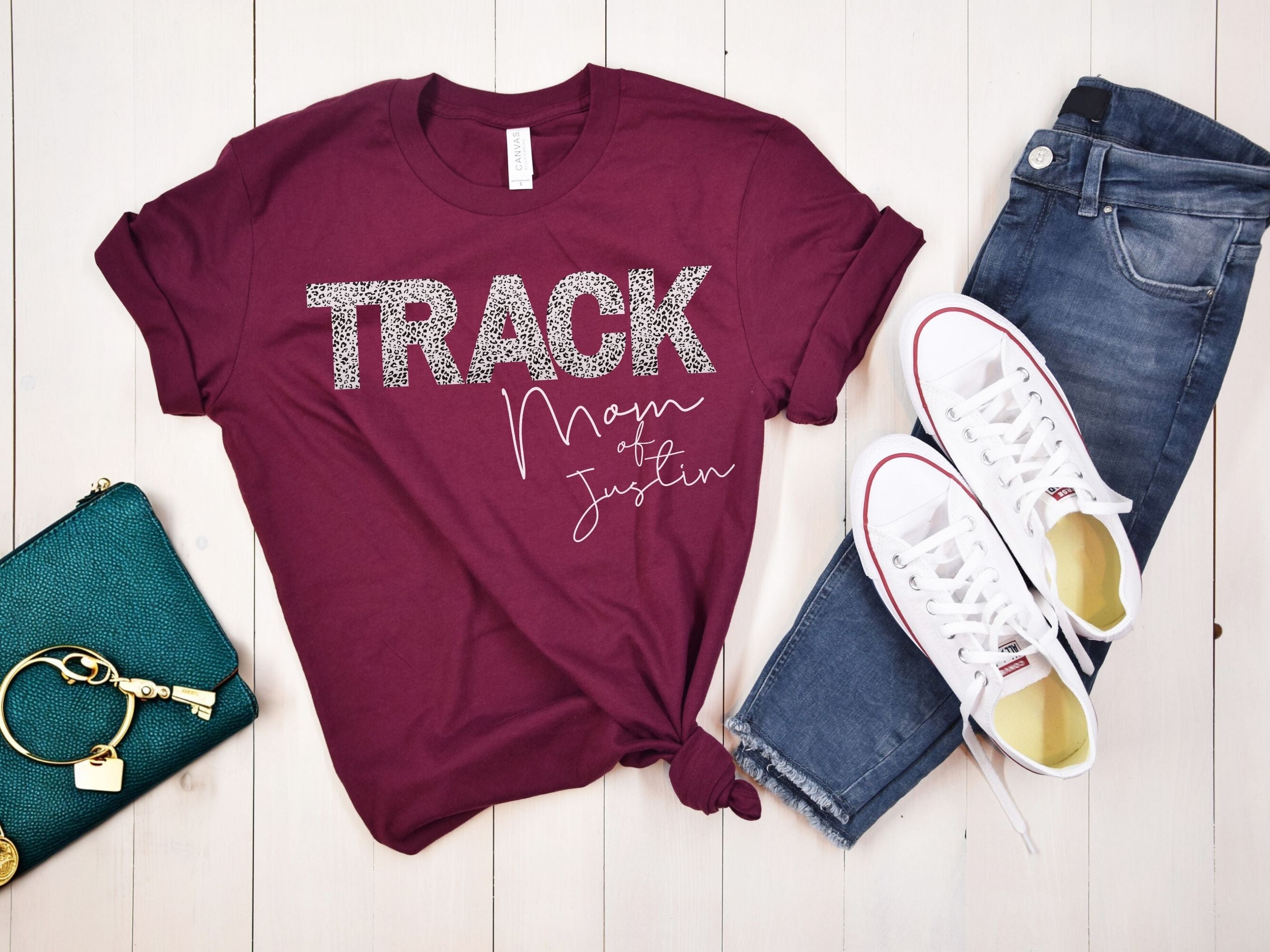 Maroon t-shirt with 'TRACK Man of Justice' text, white sneakers, blue jeans, and teal wallet on a wooden surface.