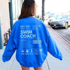 Person wearing a blue sweatshirt with 'SWIM COACH' text on the back, standing on a sidewalk.